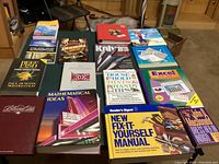 Photo showing 15 assorted books arranged on a table, titles visible include Alaska travel, reef fish, knives, household tips, Excel guide, math ideas, home repair manual, and others.