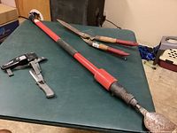 Three tools on a green surface: red-handled Nematic air concrete chipper, grey hose clamp pliers with black grips, and manual hedge clippers with wooden and red handles.