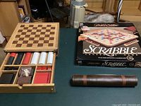 Photo showing the wooden multi game set with chess and checkers pieces, poker chips, dice, cards, the Deluxe Edition Scrabble game boxed set, and the leather roll-up game board arranged on a surface.