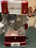 Front view of red and white Nostalgia Electrics popcorn maker, showing clear plastic front, internal popcorn kettle, two red buttons, and metal latch on the side door. Visible slight crack in plastic panel.