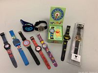 Ten children's character wristwatches arranged horizontally, showing an assortment of superhero and cartoon themes with colored bands.