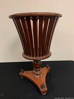 Front angled view of slatted wood planter/plant stand showing tapered basket shape and pedestal base with 3 legs and finials.