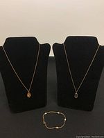 Two gold-tone necklaces with pendants displayed on black velvet busts, with a gold-tone bracelet laid flat in front.