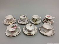 Seven bone china teacup and saucer sets arranged in two rows showing floral designs and gold trim.