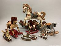 Photo showing eight vintage rocking horse figurines of various sizes and materials including wood, metal, and porcelain, arranged in a group.
