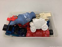 Eight vintage Avon collectible aftershave and cologne bottles in a clear plastic bin viewed from above. Multiple vehicle-shaped bottles in red, blue, and black including a white ceramic covered wagon with gold cap on top.