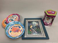 Seven Disney-themed plastic plates, a framed Cinderella picture, and a boxed Furby plush toy shown on a white surface.