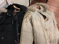 Both winter coats, one black and one beige, shown hanging on hangers on a wood floor, showing front details and fur trimmed hoods.