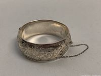 Close-up angled view of the sterling silver etched bangle with floral designs visible, along with the attached safety chain