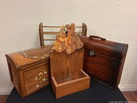 Photo of three items: wooden shoe rack with curved rails, hand-painted chest with floral designs, and wooden wall shelf with bear and doll cutout on top, all shown on black surface against wall.