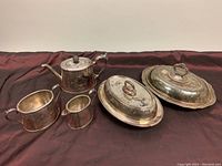 Full set of vintage silver plated servingware including teapot, creamer, sugar bowl, and two oval covered dishes on a dark fabric background.