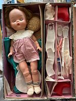 Top view of Marybel doll in original box showing doll wearing pink gingham romper with white collar and white socks. Box compartments hold red crutches, white leg casts, footwear, and other medical accessories.