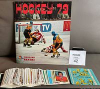 Photo showing the 1979 Panini Hockey album upright with a hockey game cover image and a spread of hockey stickers laid out in front.