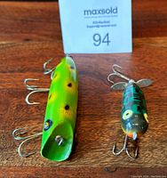 Two vintage wood fishing lures side by side on a wooden surface, green Heddon Baby Torpedo and blue-green South Bend Bass Oreno #973 with card number 94