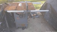 Front and top view of Delta Milwaukee table saw with rust on metal table top, central blade slot, gauge wheel for adjustments, and extension table on right side. Machine sits on gray metal frame base with sign of previous repairs and dirt buildup.