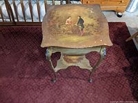 Top-down photo of a Victorian wooden table with painted figures and scene on the tabletop and lower shelf, showing worn and soiled surfaces.