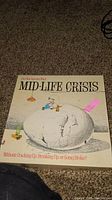 Photo of Mid-Life Crisis board game cover showing artwork of a cracked egg with a person inside, and tagline.