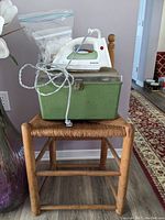 Wooden chair with woven rush seat, green sewing box on seat, Rowenta model DM-273 iron on top of sewing box with coiled power cord
