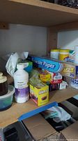 Shelf containing assorted cleaning and household supplies including freezer paper, plastic wrap, cleaner bottles, and boxes of wipes.