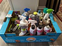 Full overview of multiple assorted cleaning and laundry bottles including carpet stain cleaners, spray bottles, and lint brush in a cardboard box