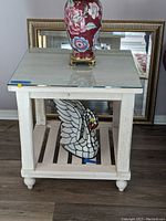 Photo of distressed wood end table with glass top showing square shape, slatted bottom shelf, and rounded legs with turned feet. Clear glass top protects surface.