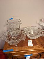 Set of three glass items on wooden surface: crystal ice bucket with handles, frosted petal-shaped bowl, and clear bowl with white floral etching and feet.