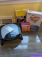 George Foreman grill with three boxed kitchen scales placed on floor against wall