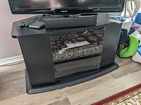 Front view of the black laminate media stand with glass door, showing scratches on top surface and a TV placed on top with remote controls on the stand.