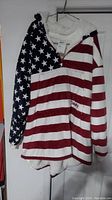 Full view of the Comfy hooded pullover featuring American flag design with stars and stripes pattern on sleeves and body, hanging on a wire hanger.
