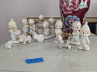 Collection of twelve figurines including Precious Moments children, Enesco figures, and one unmarked figure posed on a table with a decorative floral vase in the background.