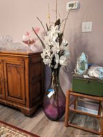 Full view of tall purple glass vase with silk white flowers and decorative branches inside, situated next to wooden furniture and other household items