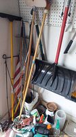 Photo showing various long-handled yard tools against a pegboard wall including rakes, shovels and a large black snow/debris shovel with a red handle, along with gardening supplies on the floor such as gloves and pots.