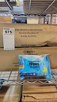 Two photos showing brown cardboard boxes labeled with SAM HEDAYA CORPORATION from Avenel, NJ, sealed and unopened. Blue packs of antibacterial hand wipes, 75% alcohol, positioned in front of the boxes for scale and content identification.