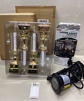 Two plastic trays holding 8 new gold-based window candles, battery covers, and bulbs. A black laser light projector and its box is on the side.