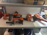 Five orange pneumatic nailers and air tools displayed on a white shelf with pegboard backing. They include various sizes of nailers suitable for framing and finish work.