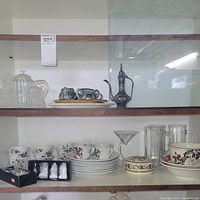 Two shelves with assorted porcelain and glassware including floral patterned tea sets, a clear glass teapot, silver salt and pepper shakers, and a decorative metal coffee pot.