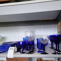 Shelf display of cobalt blue glassware including stemware, drinking glasses, and bowls with black stems and clear glass stemware present.