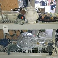 Two shelves with assorted items: ceramic teapot, glass cake stand, small glass jar, several glassware pieces including goblets and vases, jewelry in display case.