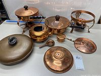 Photo showing group of copper kitchen items including bed warmer, chafing pans burners, kettle, small pans, lids, and measuring cup arranged on table.