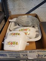 Top-down view of the Corning Ware Spice of Life bakeware in cardboard box showing four white ceramic pieces with glass lids and handles with colorful vegetable design.