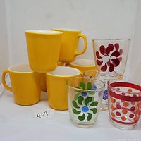 Photo of 6 solid yellow Corning/Corelle mugs stacked and 4 vintage sour cream glasses with colorful retro designs including red, green, blue, and orange patterns.