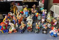 Wide shot displaying a group of 28 various clown figurines placed on a table. The figures vary in size, posture and costume style, mostly colorful and detailed resin clowns including some seated in small cars and some holding balloons.