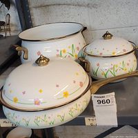 Front view of three Lincoware metal cookware pieces with wildflower design: a large pot without lid, a covered saucepan, and a covered sauté pan, all with white and floral exterior and gold-toned handles and knobs.