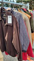Photo showing seven men's jackets on hangers outdoors, including brown, gray quilted, red, tan, and other colored jackets lined up on a rack.