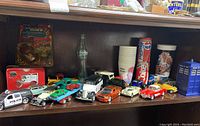 Shelf displaying multiple vintage toy cars in various colors and designs, including police and sports cars, with a vintage glass bottle and branded cups visible