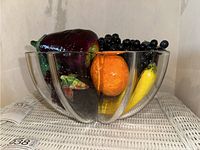 Glass bowl filled with various faux fruit including purple eggplant, orange tangerine, yellow banana, dark red bell pepper and cluster of black grapes.