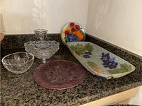 Five items on black granite countertop: clear cut glass vase, footed candy dish, medium bowl, large colorful hand painted fruit platter, small hand painted fruit round plate on stand.