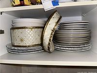 Stack of plates and a large decorative bowl with diamond quilted pattern and gold accents inside a cabinet shelf.