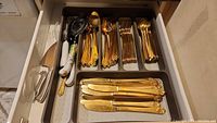 Dresser drawer with multiple gold tone forks and spoons arranged in a tray, plus knives with floral ceramic handles.
