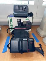 Front view of Polaroid Impulse camera on top of original box and Nikon TeleTouch 300 AF camera in front with carrying case behind it.